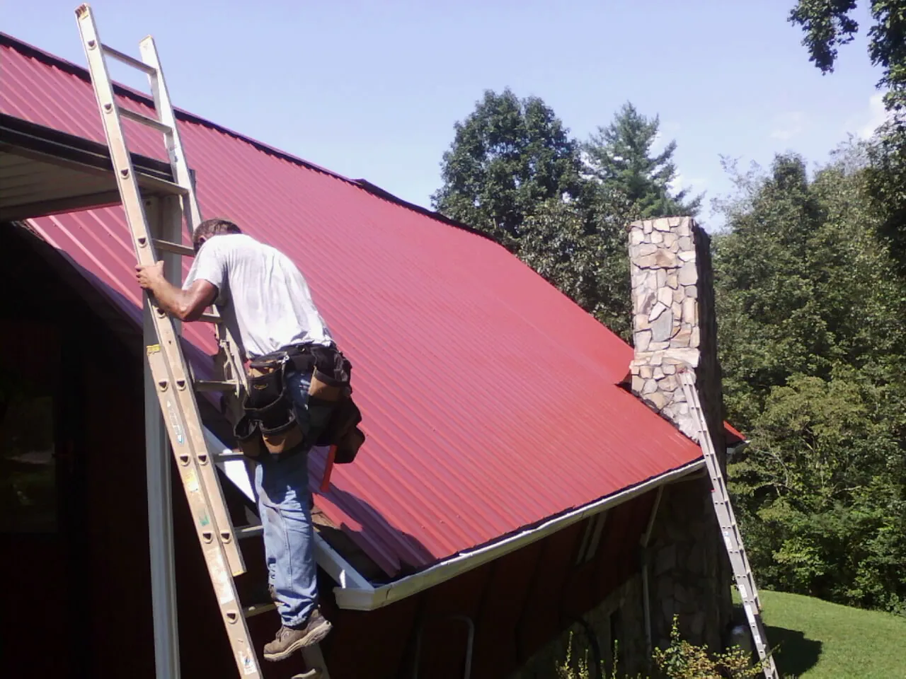 Roofer on a ladder beside a red metal roof and stone chimney — Soffit & Fascia Repair by Roofing Brothers in Little York